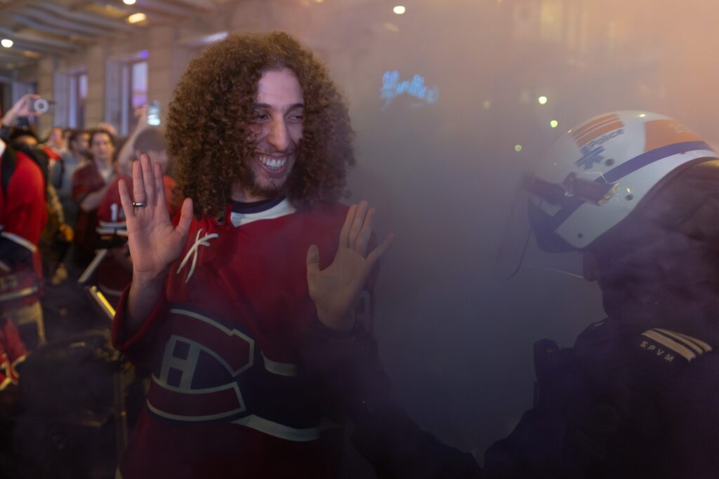 A man in a Canadiens jersey smiles at a police officer in riot gear as they're surrounded by smoke