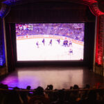 People sitting in movie theatre watching hockey game on big screen.